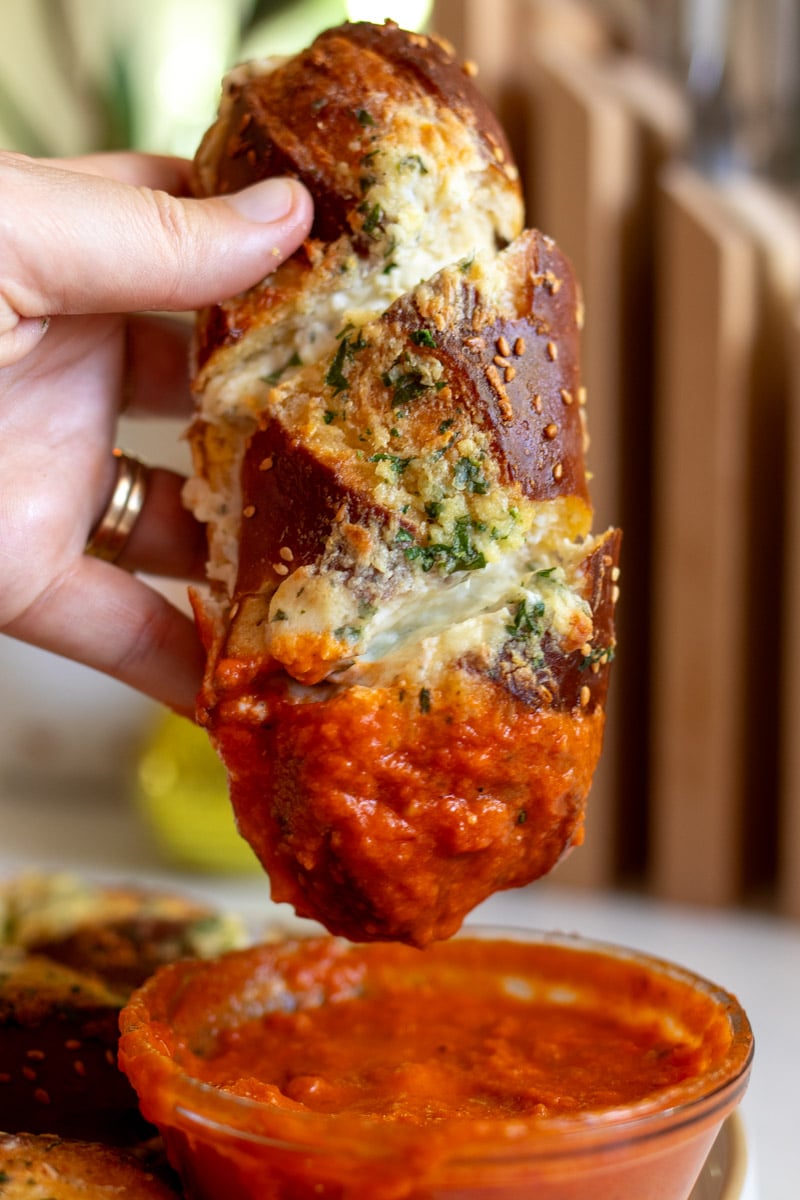 a piece of maurice's cheesy garlic pretzel bread being held over a container of marinara sauce.