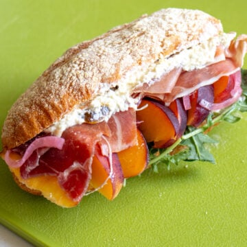 a prosciutto, peach and feta sandwich on a green cutting board.