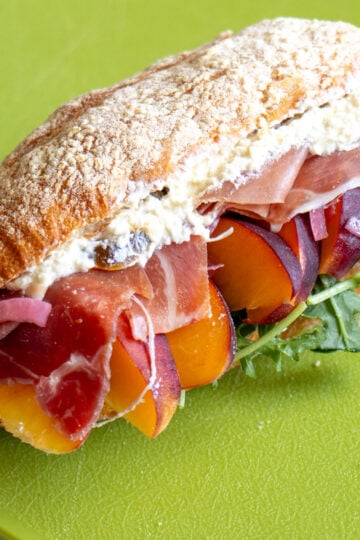 a prosciutto, peach and feta sandwich on a green cutting board.