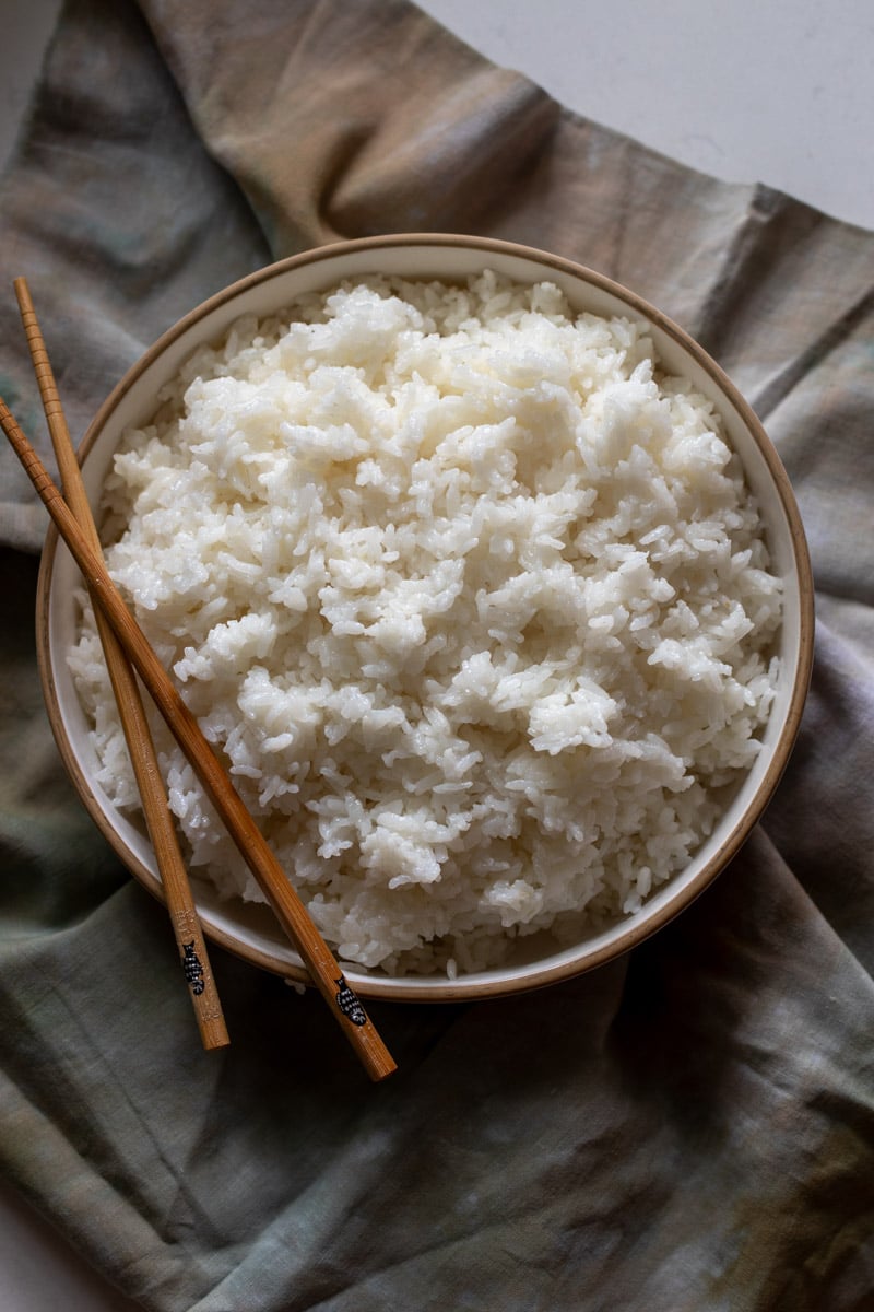 a white bowl of sushi rice with wooden chopsticks on the side on top of a dark napkin.