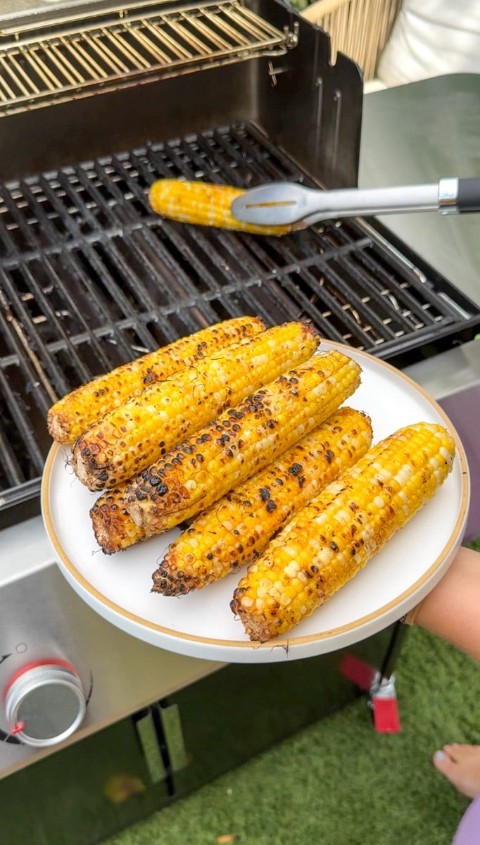 a few ears of corn on a plate with the grill in the background still grilling a corncob.