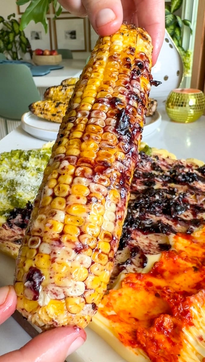 a grilled corn cob rolled in blueberry lemon butter with a colorful butter board in the background.
