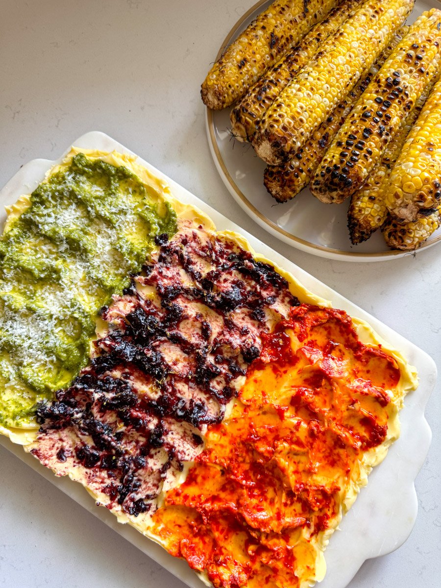a colorful butter board next to a plate full of grilled corn cobs.