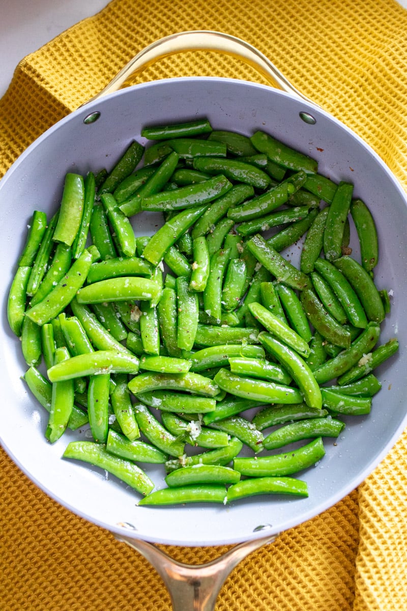 an overhead view of a white carway frying pan with sugar snap peas being sauteed. 