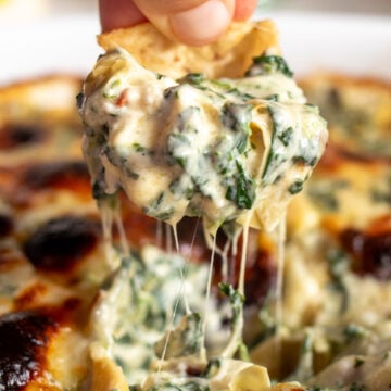 a chip being dipped in ultra cheesy spinach artichoke dip with a cheese pull coming out of it.