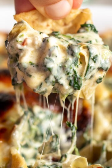 a chip being dipped in ultra cheesy spinach artichoke dip with a cheese pull coming out of it.