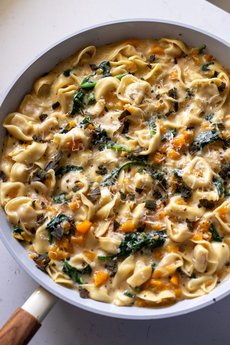 a big white frying pan filled with cheese tortellini that are coated in a creamy sauce with bits of butternut squash, spinach and parmesan.