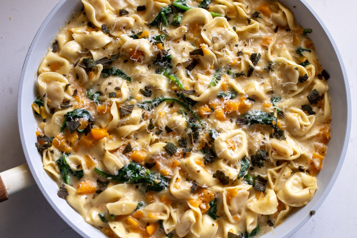 a big white frying pan filled with cheese tortellini that are coated in a creamy sauce with bits of butternut squash, spinach and parmesan.