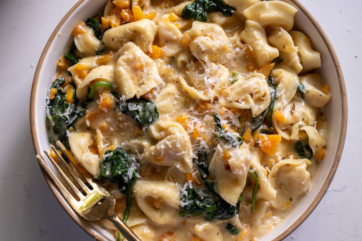 a white bowl filled with creamy tortellini in a brown butter sauce with fall veggies.