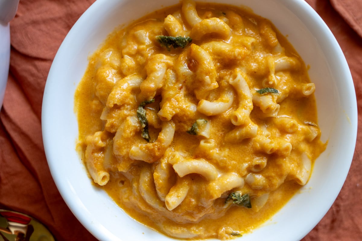 a white bowl full of creamy orange pumpkin macaroni and cheese with bits of sage on top and a burnt orange napkin underneath.