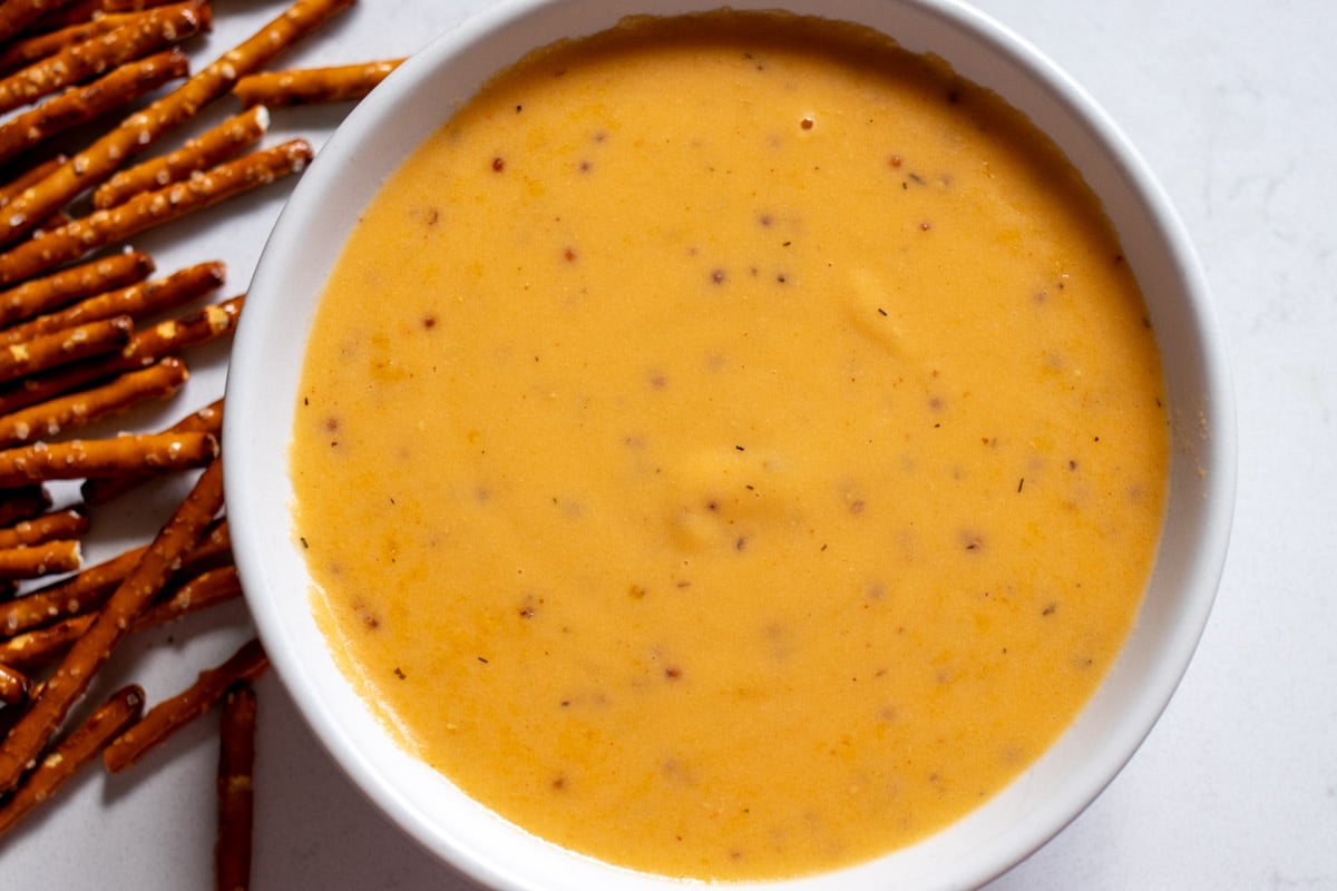 a white bowl of beer cheese dip specked with grainy mustard with a side of pretzels.