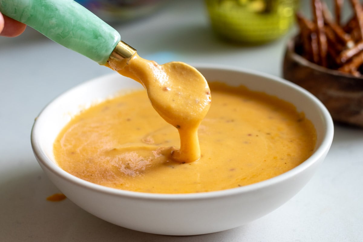 a white bowl full of creamy beer cheese dip made with cheddar and lager with a green spoon scooping up a spoonful.