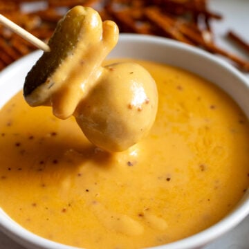a pickle, pretzel, potato skewer being dipped into creamy beer cheese dip that's made with cheddar and lager.