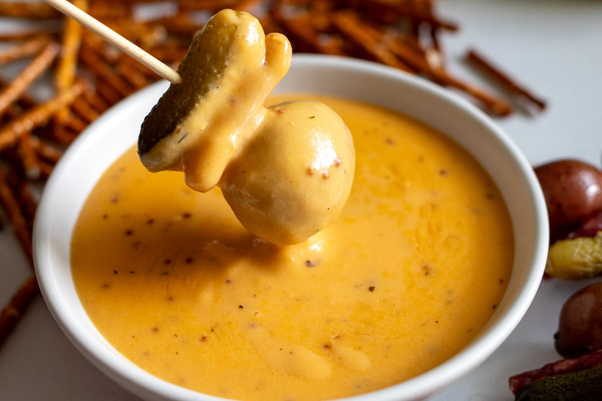 a pickle, pretzel, potato skewer being dipped into creamy beer cheese dip that's made with cheddar and lager.