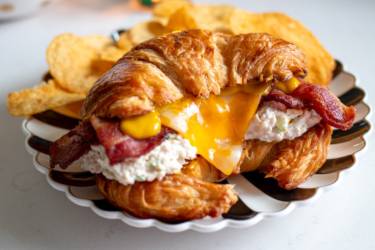 a chicken salad croissant sitting on a black and white plate with chips behind it.