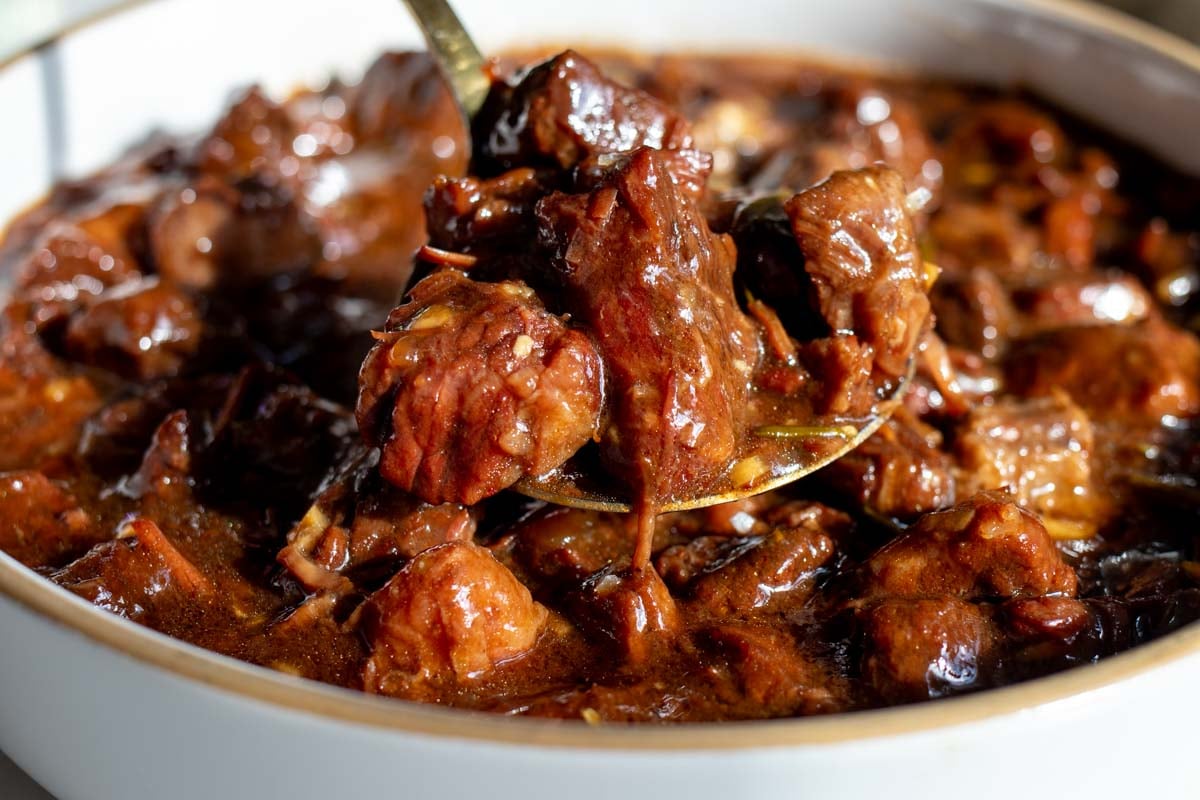 a spoon holding a scoop of tender beef bites in a rich gravy with bits of fresh rosemary in it.