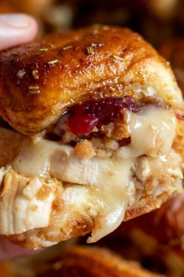 a hand holding a thanksgiving inspired turkey cranberry slider with brie cheese dripping from it and stuffing coming out.