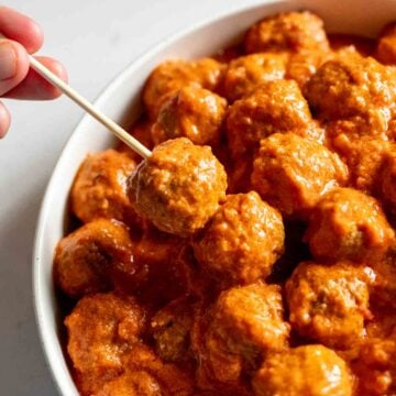 a hand holding a toothpic with crockpot cooked meatballs in a creamy tomato sauce.