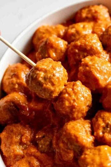 a hand holding a toothpic with crockpot cooked meatballs in a creamy tomato sauce.
