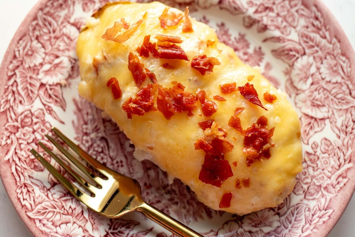 a single cheesy twice baked stuffed potato with crispy bits of prosciutto on top with a gold fork and pink and white plate.