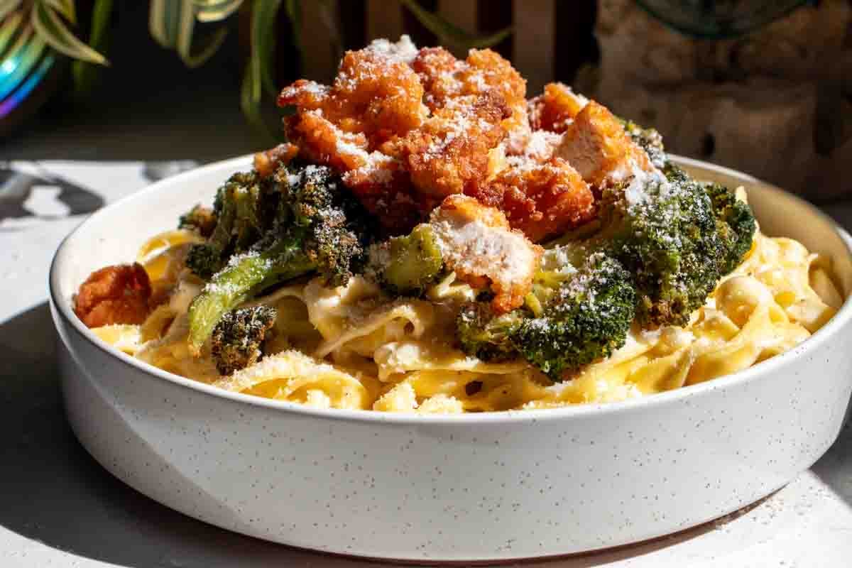 a white bowl full of crispy chicken, roasted broccoli and creamy fettuccine alfredo with a sprinkle of parmesan on top.