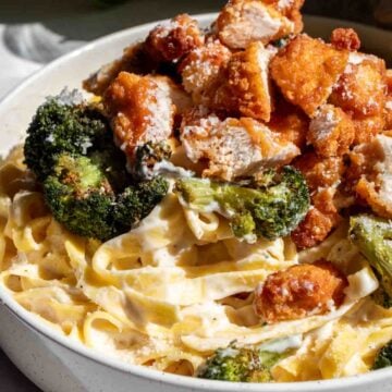 a white bowl full of crispy chicken, roasted broccoli and creamy fettuccine alfredo with a sprinkle of parmesan on top.