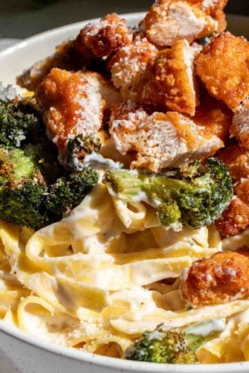 a white bowl full of crispy chicken, roasted broccoli and creamy fettuccine alfredo with a sprinkle of parmesan on top.