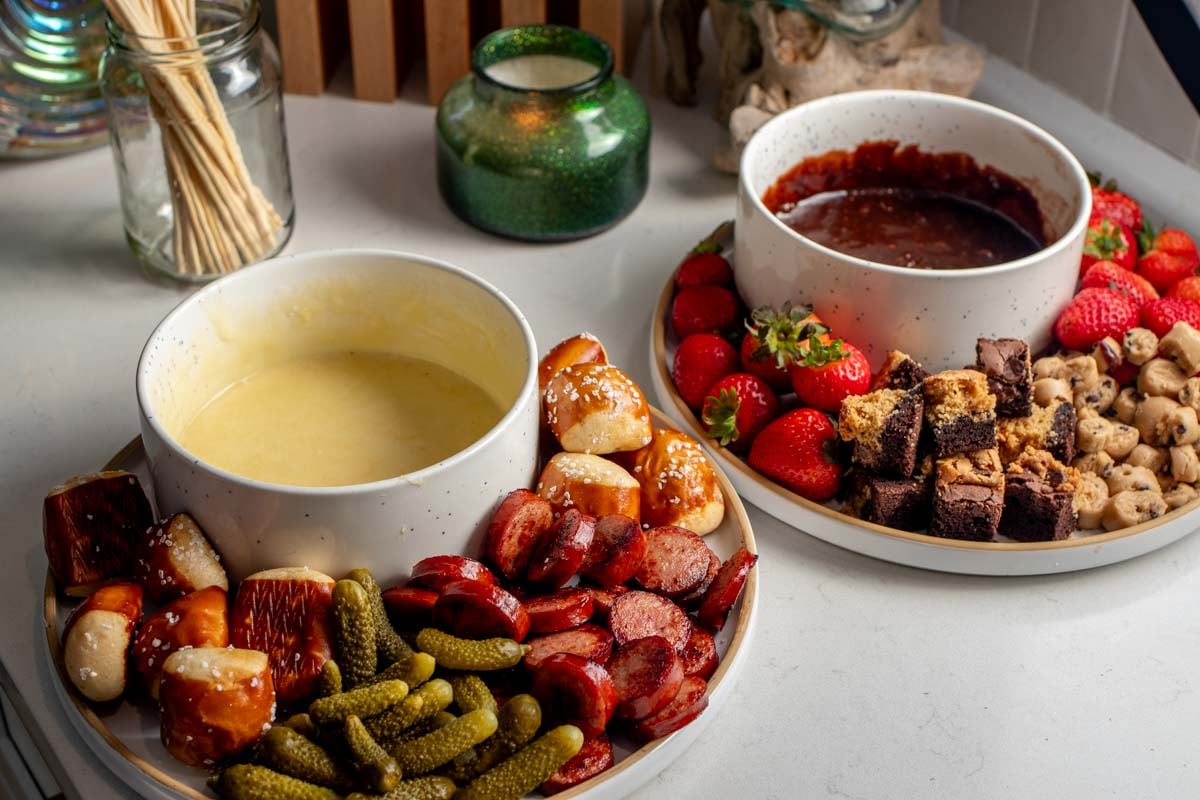 two fondue sets on a table with colorful dippers like fruit, crackers, pickles, sausage and more. 