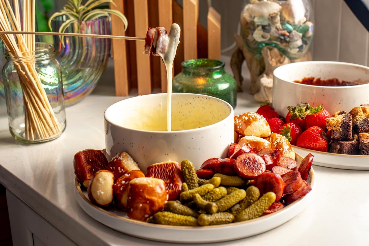 melted cheese fondue in a white bowl with a fondue fork, pickle, and other colorful dippers. 