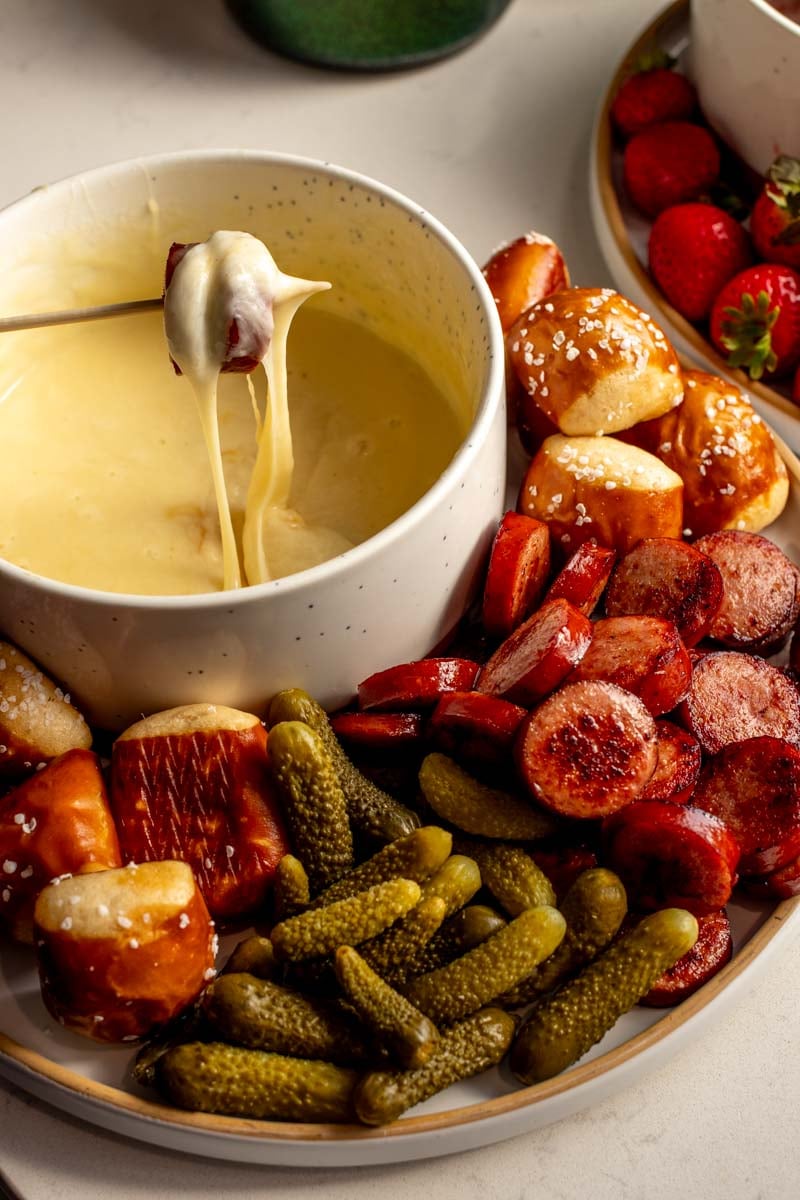 a fondue fork holding a pickle, sausage and a cheese pull.