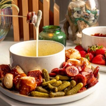 a bowl of cheese fondue with a fondue fork holding up some melty cheese and sausage. it's also got other vegetables, pickles, and bread shown on a serving plate.