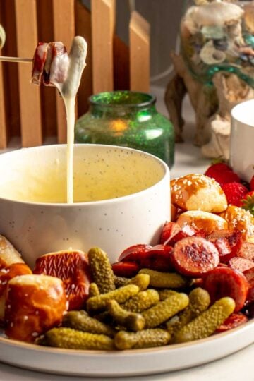 a bowl of cheese fondue with a fondue fork holding up some melty cheese and sausage. it's also got other vegetables, pickles, and bread shown on a serving plate.
