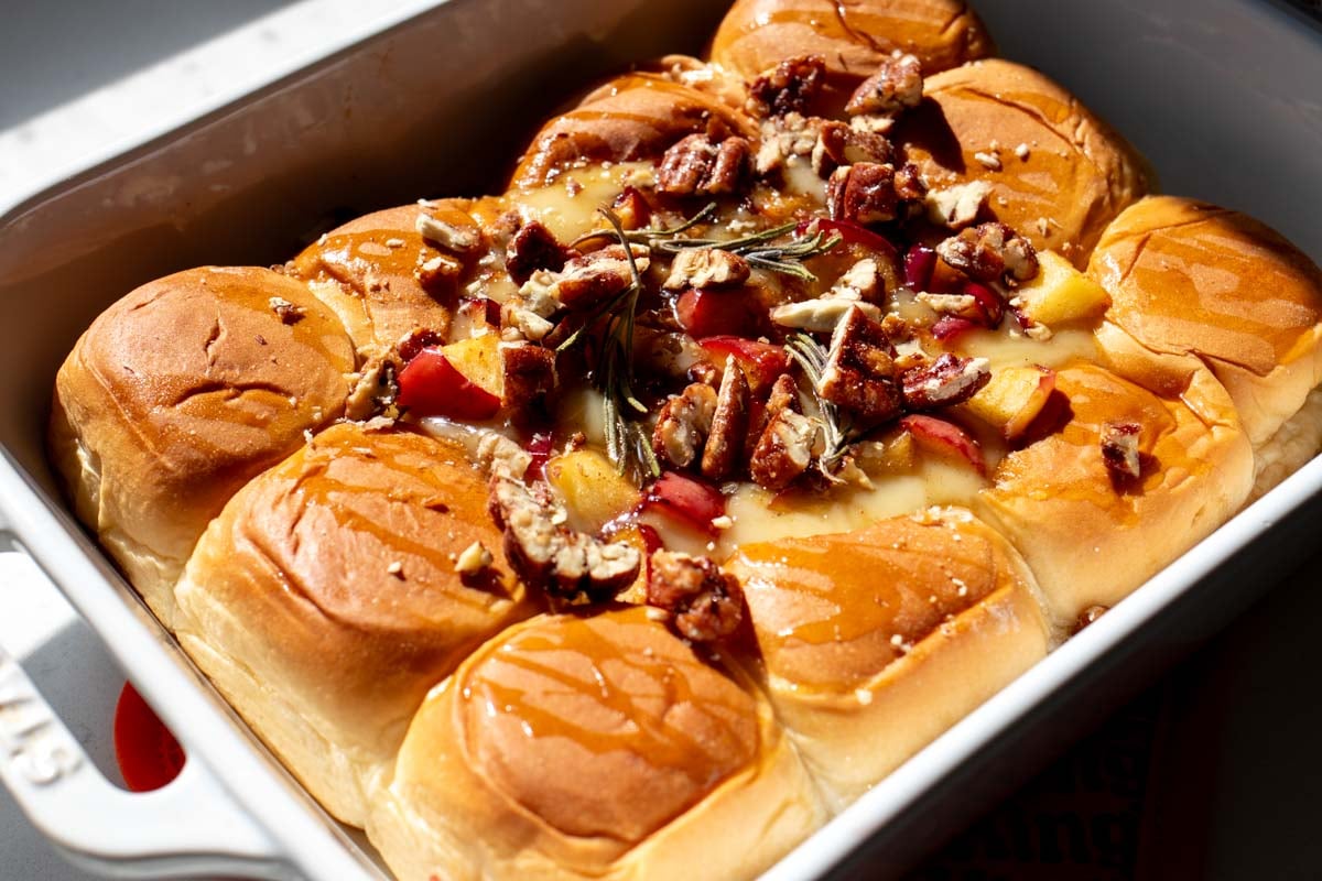 a white baking dish filled with glazed hawaiian rolls, melty brie, and caramelized apples.