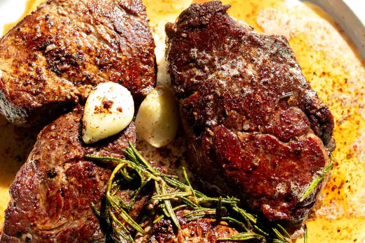 an up close look at some perfectly cooked filet mignons with green rosemary and garlic cloves on a white plate.