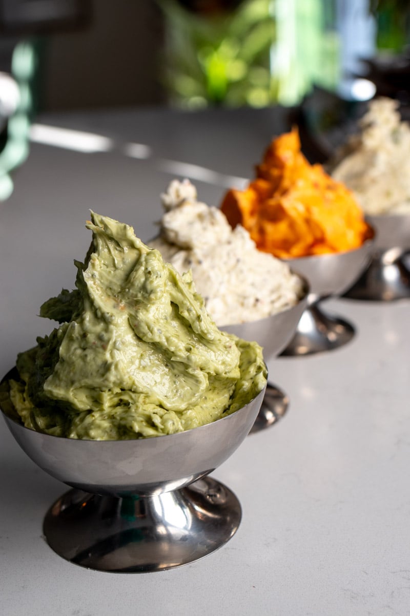 a collection of colorful steak butters in silver dishes on a white countertop.