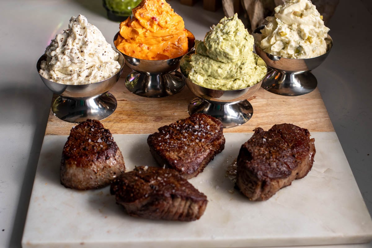 4 colorful compound butters for steak in silver cups with 4 perfectly seared steaks on the side.