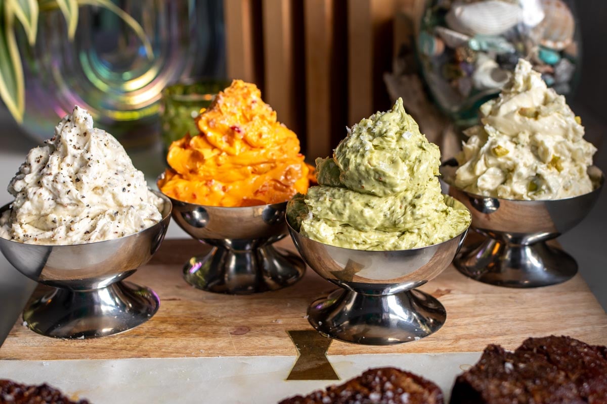 4 colorful compound steak butters in silver dishes lined up on a cutting board.