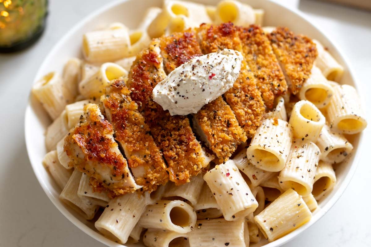 a white bowl full of creamy cacio e pepe pasta with a crispy chicken cutlet on top.