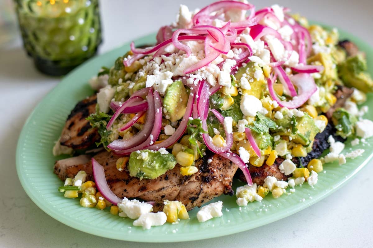 grilled chicken with sear marks on a light green plate with a colorful avocado corn feta salad topped with pickled red onions.