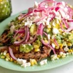 grilled chicken with sear marks on a light green plate with a colorful avocado corn feta salad topped with pickled red onions.