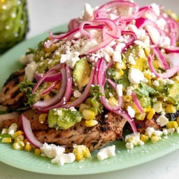 grilled chicken with sear marks on a light green plate with a colorful avocado corn feta salad topped with pickled red onions.