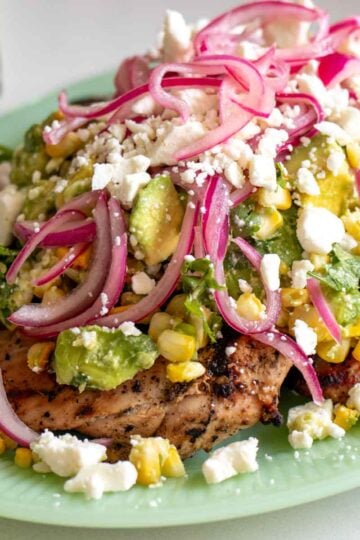 grilled chicken with sear marks on a light green plate with a colorful avocado corn feta salad topped with pickled red onions.