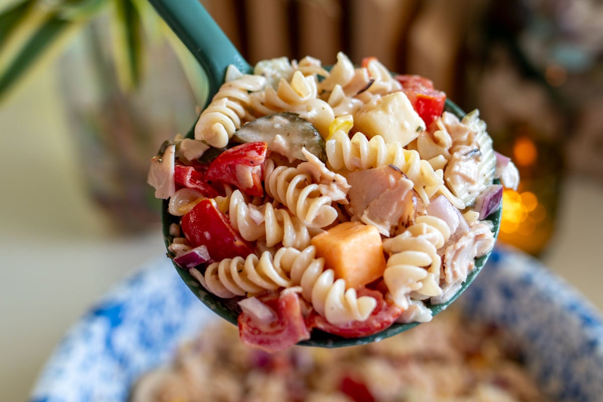 a giant serving spoon holding a scoop of the sub in a tub pasta salad with some greenery in the background.