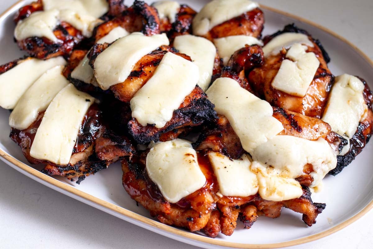 bbq chicken thighs on a white platter with lots of white melted cheese on top.