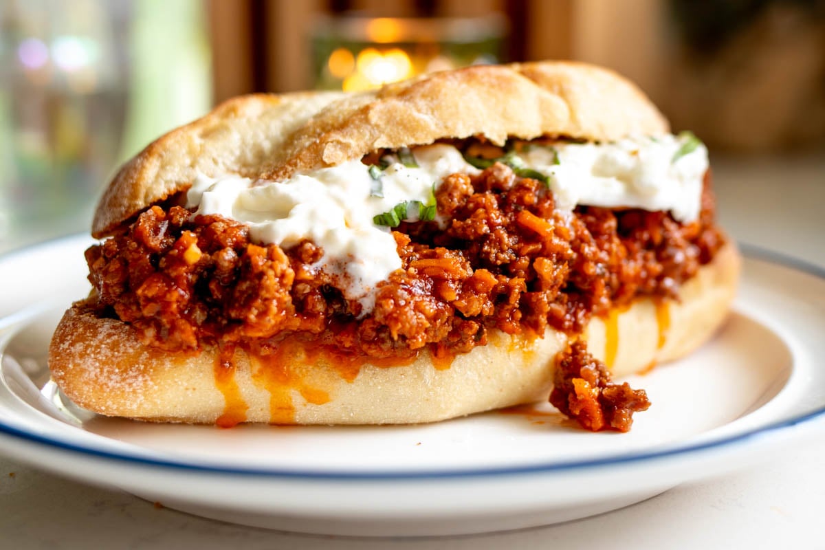 a close up of a big sandwich made of toasted french bread and a bright red italian sloppy joe mixture with globs of burrata cheese and bits of green basil on a white plate.