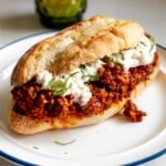 a big sandwich made of toasted french bread and a bright red italian sloppy joe mixture with globs of burrata cheese and bits of green basil on a white plate.