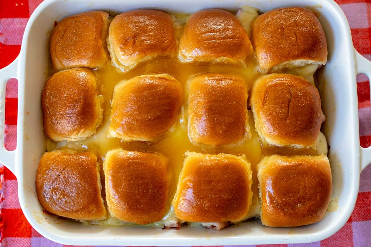 a white baking dish full of glazed ham and cheese sliders with a pink and red plaid cloth underneath.
