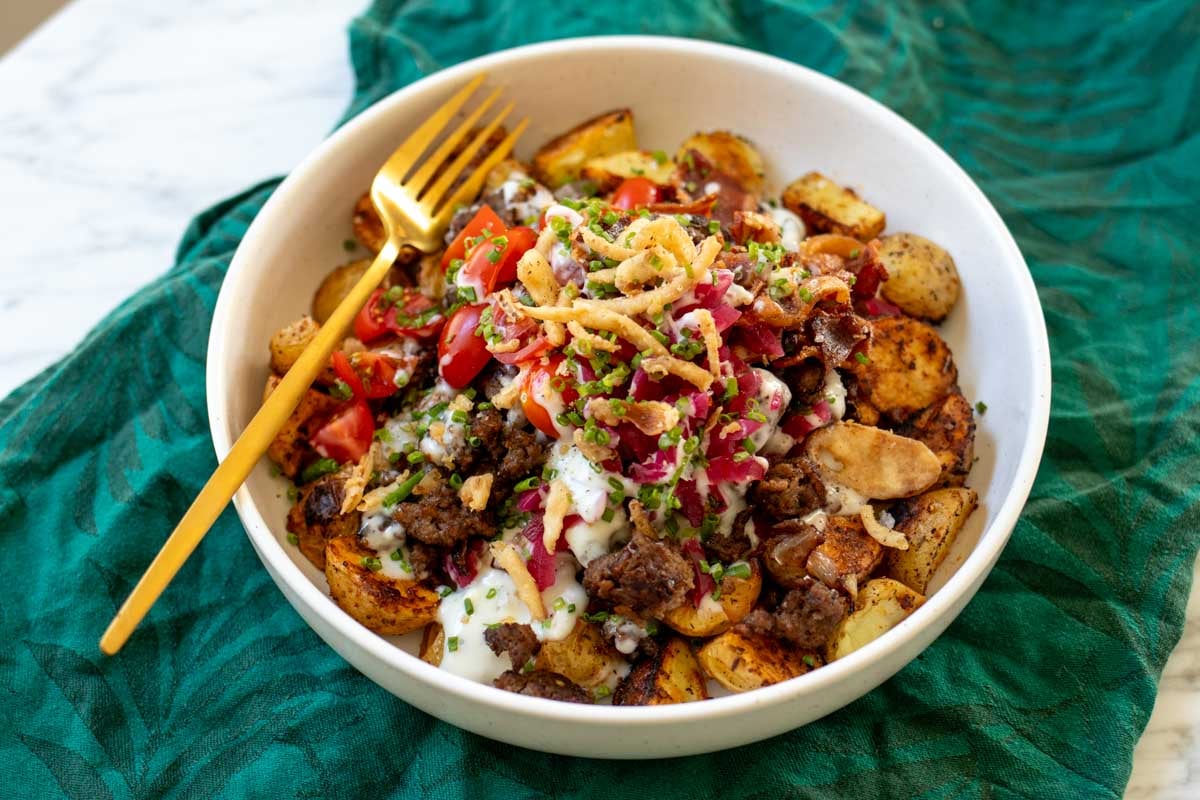 a white bowl full of roasted potatoes, ground beef, colorful toppings on a marble countertop with a green napkin.