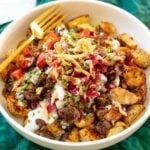 a burger bowl with potatoes, ground beef, toppings, a gold fork and a green cloth underneath.