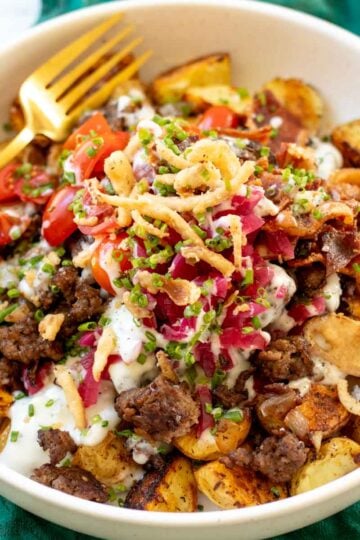 a burger bowl with potatoes, ground beef, toppings, a gold fork and a green cloth underneath.